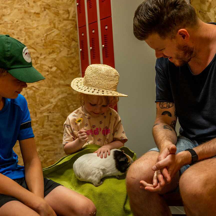 Meisje met rieten hoed aait een cavia die bij haar op schoot zit. Man en jongen kijken toe